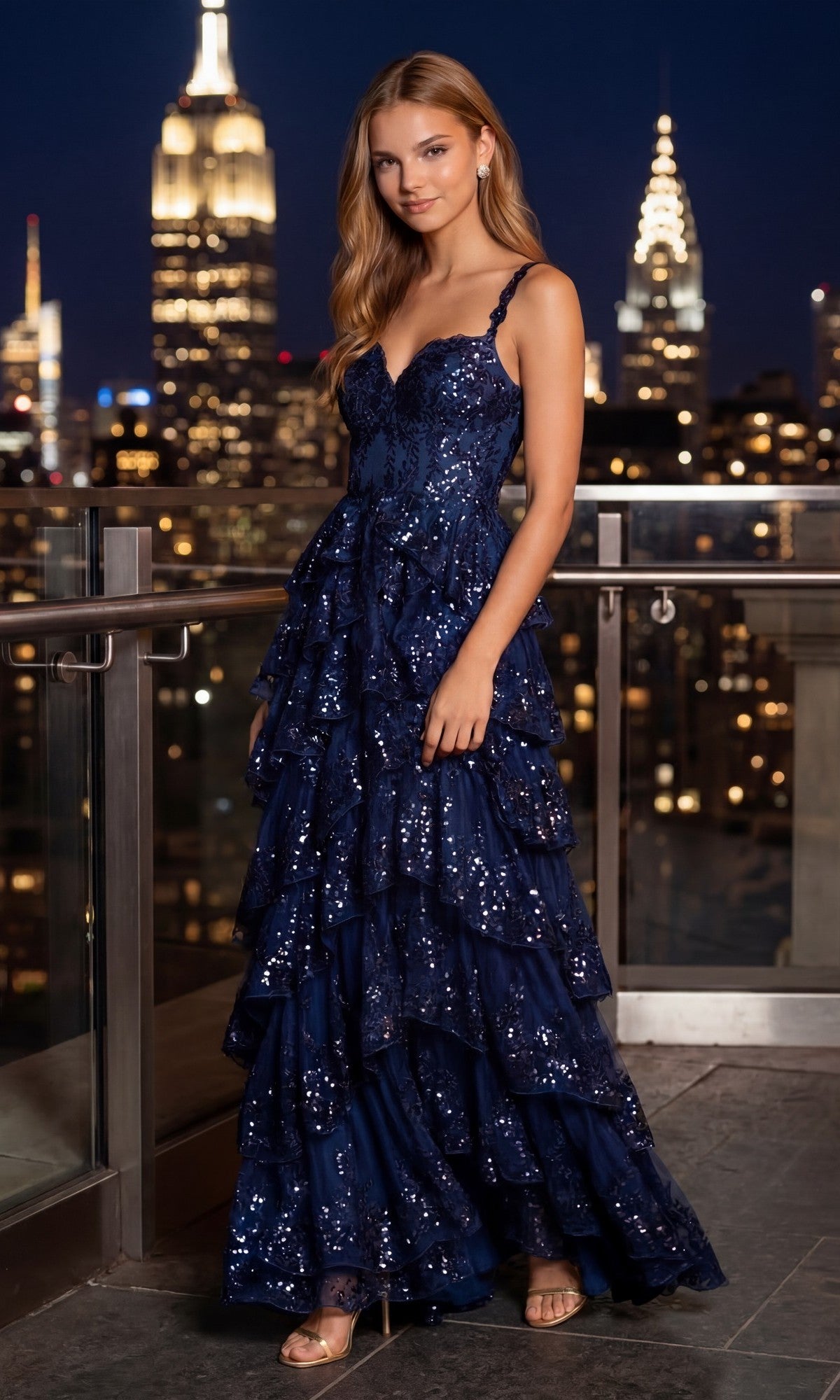 A woman in a navy Beaded-Lace Ruffle Prom Gown (Blondie Nites 5394BN) with ruffled tiers and a corset-style bodice stands on a balcony at night, framed by the sparkling city skyline.